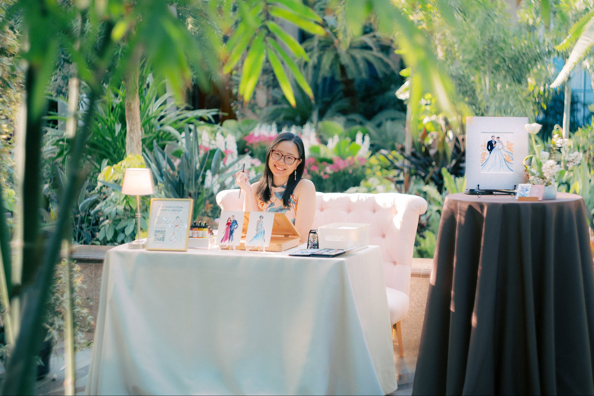 An live event artist sits behind a table in a garden greenhouse setting, on a pink chair. To the right, a matted original painting of the wedding couple is displayed on a hightop table with dark drapes. Lush green foliage frames the scene.